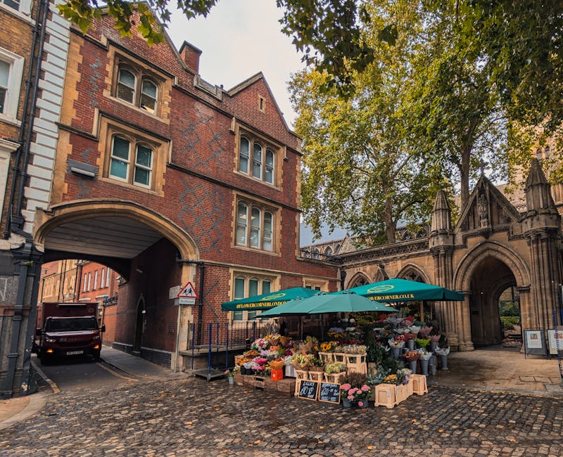 A vibrant flower stall situated on a cobblestone street near an arched stone entrance and historic red-brick buildings. The stall is shaded by a large green parasol with the label 'Floral London' and