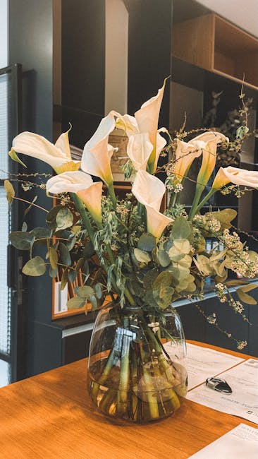 A large, elegant floral arrangement featuring cream-colored calla lilies with gracefully curved petals and long stems, complemented by sprigs of delicate baby's breath and surrounding eucalyptus leave