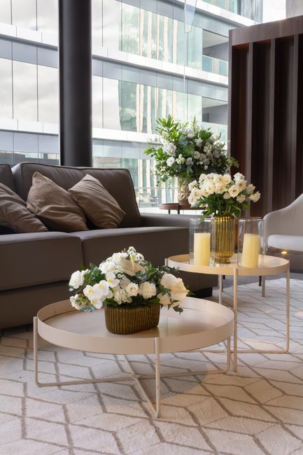 A contemporary indoor setting featuring two round, cream-colored side tables with slender metal legs, each topped with floral arrangements. The front table displays a basket-style arrangement of predo
