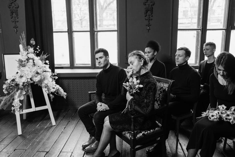 A black and white photograph depicting a group of six individuals seated indoors during a funeral ceremony, arranged in front of large windows that allow natural light to illuminate the scene. The for