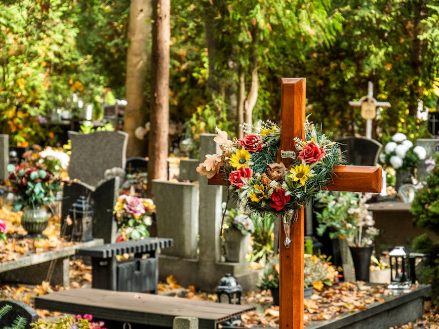 A wooden funeral cross decorated with a floral arrangement consisting of red roses, yellow daisies, and beige flowers, surrounded by lush green foliage. The cross is positioned among other memorial st