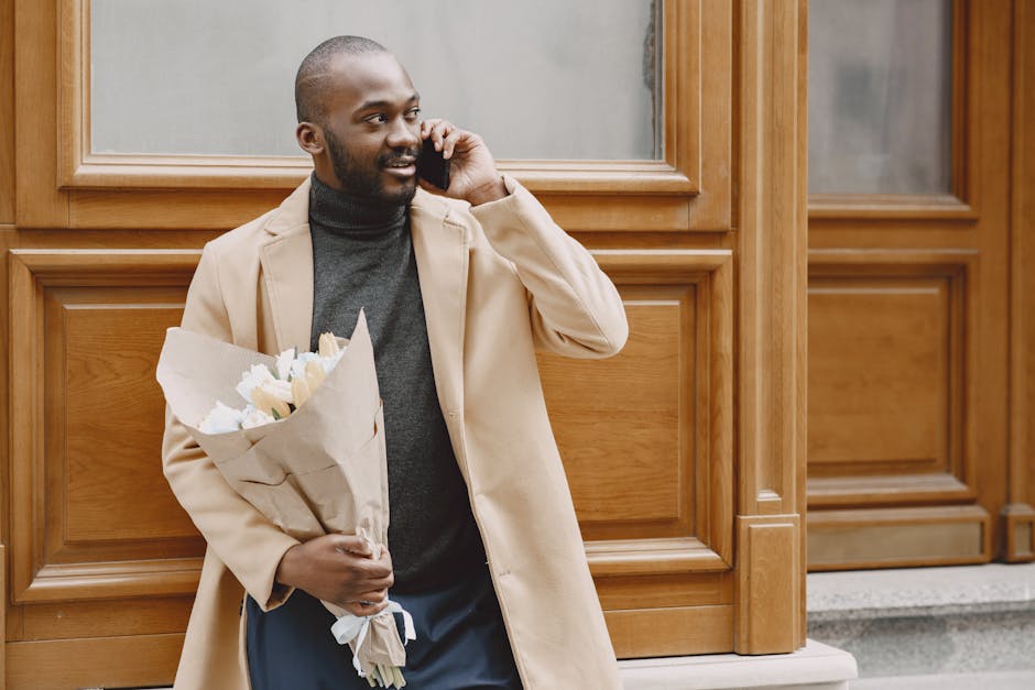A young man with dark skin, a closely shaved head, and a well-groomed beard is smiling while talking on his mobile phone. He is standing outdoors in front of a wooden-paneled building with large, fram