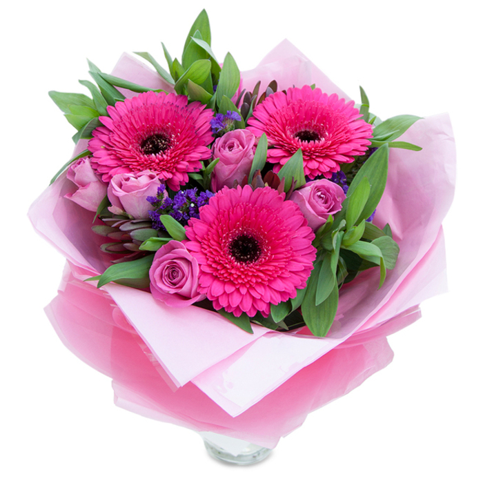 A lively trio of fuchsia Gerbera daisies sits at the centre of this arrangement, each bloom revealing rows of saturated petals around a boldly dark eye that gives a graphic, celebratory feel. Surrounding them are roses in a soft lavender-pink, their petals held in fresh, budding formation and lending a romantic, almost whisper-like softness to the overall colour story. Interspersed throughout are tiny bunches of purple statice-a crisp, papery texture that introduces cool tonal contrast and a meadow-like lightness. The foliage is generous and green, with broad leaves that read as salal and occasional elongated eucalyptus leaves, their crisp edges and matte sheen framing the composition and amplifying the pink hues. The bouquet is cocooned in an inner layer of matte pale pink tissue paper, which supports the stems and cushions the blooms; over this, a sheet of translucent fuchsia-pink cellophane, slightly crinkled, wraps the stems with a subtle shimmer that catches morning light and gives the presentation a polished, gift-ready finish. The florist's folds are visible and neat, and the lower edge reveals the top rim of a clear glass vase, suggesting the flowers are prepared for immediate display on a mantle or dining table. Photographed against a pure white backdrop, the arrangement reads as fresh, cheerful and intimate-the kind of bouquet one might send across Earls Court to celebrate a birthday, an anniversary, or to brighten a friend's flat near Warwick Road or Old Brompton Road. The scent is quietly pleasant-roses offering a soft, classic perfume while the Gerberas lend a faint, green sweetness-together creating a bouquet meant to comfort, celebrate and make someone feel noticed.